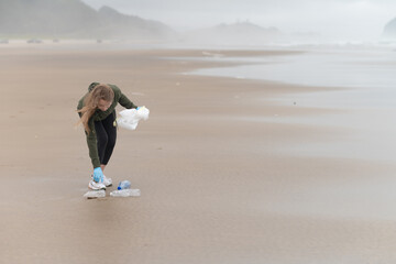 Girl volunteer with long blond hair collects trash and plastic bottles on the sandy beach. Defocused background. Environmental protection, recycling and waste disposal.