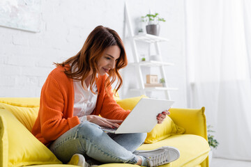 cheerful freelancer sitting on yellow sofa with crossed legs and working on laptop