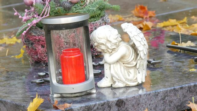 Red memorial candle burning with fire on rainy day on tombstone in cemetery autumn, rainy day. Grave with tombstone on raindrops falling, against abackground of yellow foliage next to sad angel statue