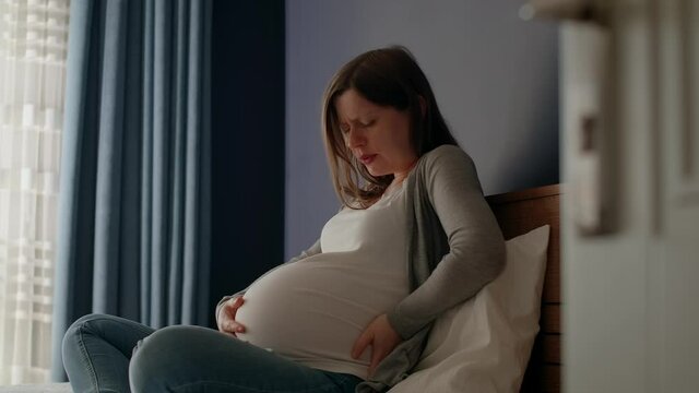 Pregnant woman with big belly sitting in the bedroom, contractions begin, she feels pain. Last month's pregnancy, starting of childbirth.