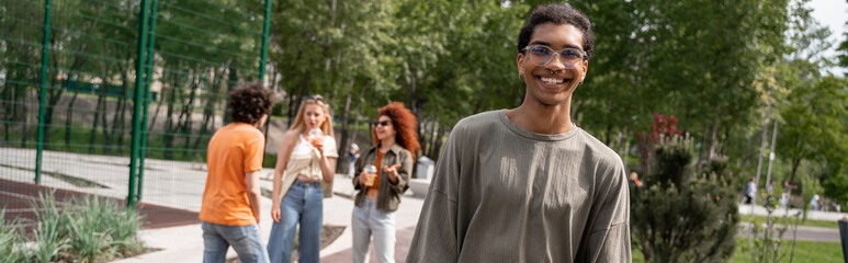 Fototapeta premium happy african american man looking at camera near friends in park, banner.