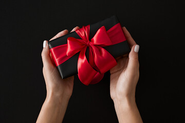 Top view photo of female hands holding black gift present with red satin ribbons on black background.