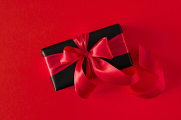 Top view photo of black gift present with red satin ribbons and bow on red background.