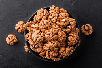 Peeled walnut in a bowl and a few nuts scattered around on a black textured background, top view.