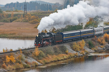 Fototapeta na wymiar Retro steam train moves along the lake at autumn cloudy time. Karelia. Russia.
