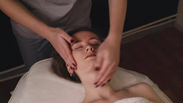 Woman masseur therapist kneading healthy head face massage relaxing professional therapy hands oil. Girl lying couch table resting gets rubdown spa salon body care concept, slow motion treatment

