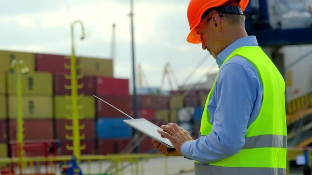 Ship cargo loading process. Grey haired general manager in bright vest fills form on tablet while workers walk past metal containers in dockyard
