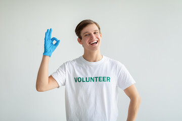 Cheerful young volunteer guy in white t-shirt and gloves showing OK gesture and smiling at camera