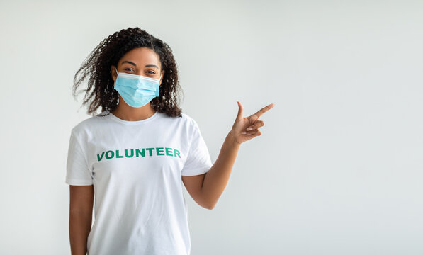 Look There. Young Female Black Volunteer In Medical Mask Pointing Finger Aside Showing Copy Space Over Light Wall