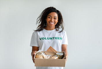 Happy black female volunteer holding donations box with cans and packages, smiling at camera,...