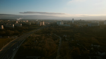 The suburb is backlit by the setting sun. Houses, a park area and a city highway are visible. Aerial photography.