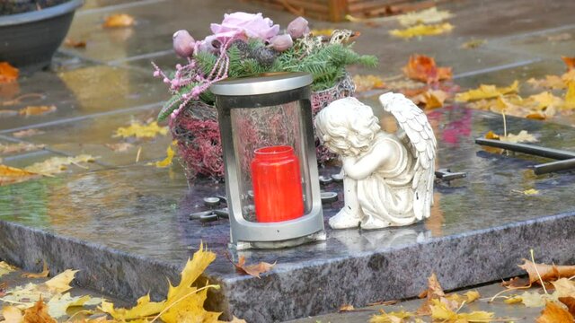 Red memorial candle burning with fire on rainy day on tombstone in cemetery autumn, rainy day. Grave with tombstone on raindrops falling, against abackground of yellow foliage next to sad angel statue