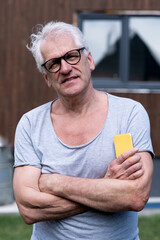 A gray-haired man with glasses smiles at the camera holding a phone against the background of a suburban wooden house on a sunny day