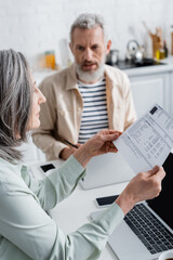 Mature woman holding paper with bills near devices and blurred husband in kitchen.