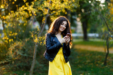 Closeup portrait of a brunette with wavy hair, wearing a leather jacket in the park, read a message on a her cell phone.