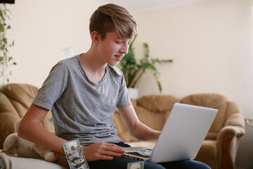 freelance teenager recounts money earned online on laptop and happy with earnings. boy studies online and earns money from comfort of home. child counting money showing it friends on conference call.