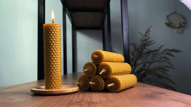 Stylish handmade candle burns on wooden tray near pile of items with honeycomb texture on rustic table in room closeup