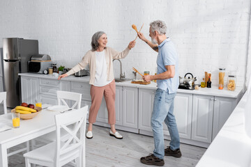 Happy mature woman fighting with wooden spatulas in kitchen.