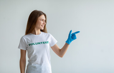Look here. Young female volunteer in gloves pointing and looking aside at free space, standing over light studio wall