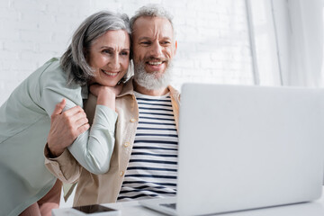 Positive mature woman hugging husband and looking at blurred laptop at home.