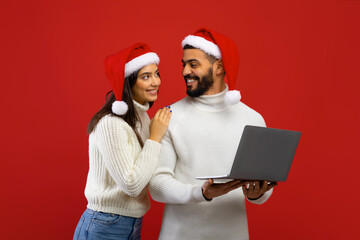 Christmas promo concept. Loving arab couple using laptop computer and smiling to each other, standing on red background