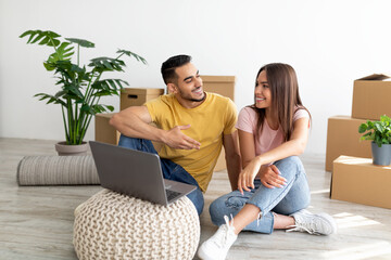 Multiracial couple with laptop buying household goods for their new house online, sitting on floor...