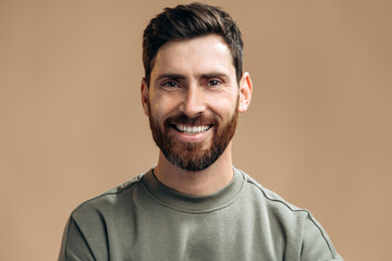Portrait of cheerful man with dark hair and beard standing and looking at the camera with satisfied face and smiling. Indoor studio shot, isolated on brown background