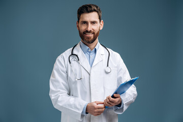 Portrait photo of handsome family doctor wearing white lab coat and stethoscope smiling friendly and posing with his folder isolated blue color background