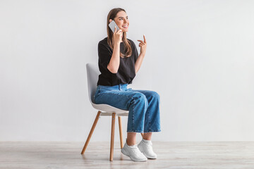 Smiling young woman having conversation on cellphone, sitting on cozy chair against white studio wall, full length