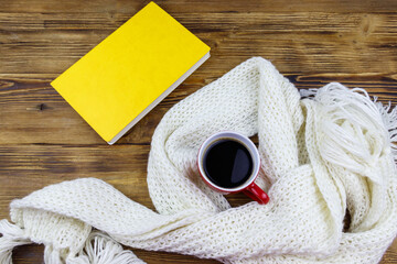 Knitted white scarf, book and cup of coffee on wooden table. Top view. Winter cozy concept