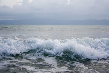 Sea wave below cloudy sky with mountains silhouette on the background