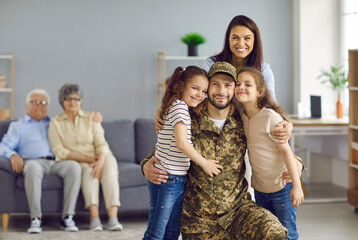 Veteran soldier comes back to his family from the military. Happy wife and two children who missed daddy a lot hugging him and looking at camera on blurred copy space home background with grandparents