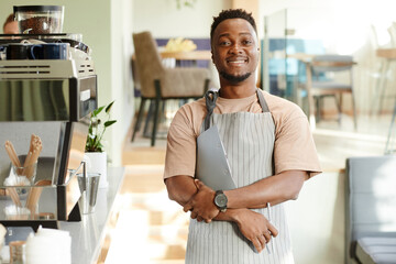 Horizontal medium portrait of joyful African American man wearing apron working in cafe holding pen and clipboard with papers smiling at camera