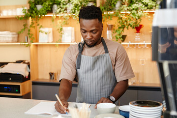 Horizontal medium portrait of young Black man wearing apron standing at cafe kitchen counter writing something in notebook