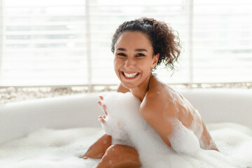 Portrait of cheerful young woman taking bath with foam, smiling at camera, relaxing in hot bathtub...