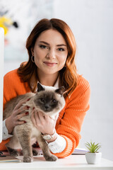 pretty woman smiling at camera while cuddling cat sitting on work desk