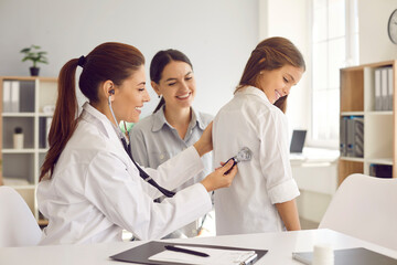 Fototapeta premium Female doctor puts stethoscope on girl's back and listens to heartbeat and lungs. Pediatrician examines teenage girl in presence of her mother. Concept of health care and pediatric medical examination