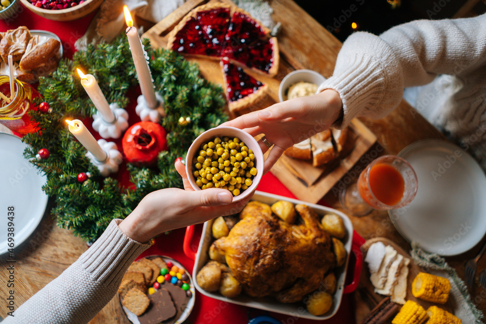 Wall mural Close-up high-angle view of unrecognizable young woman and man passing delicious food sitting at festive Christmas table during holiday family friendly party, selective focus.