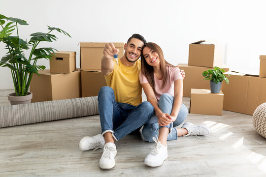 Full length portrait of loving interracial couple sitting on floor among cardboard boxes, showing house key indoors