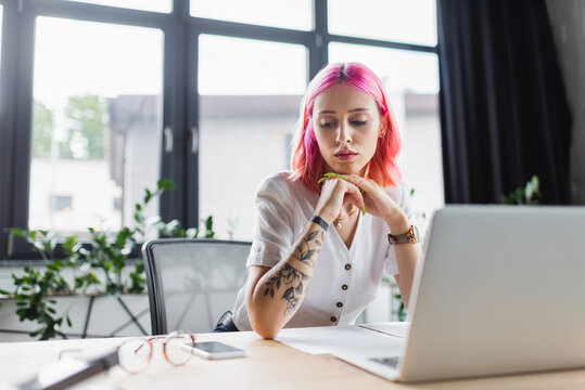 businesswoman with pink hair looking at smartphone near laptop on desk.