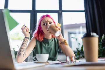 shocked businesswoman with pink hair drinking coffee near laptop in office.
