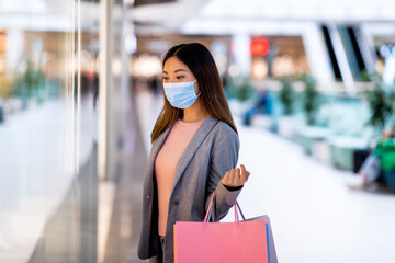 Millennial Asian woman in protective mask holding paper bags, window shopping, looking at new clothes at huge mall