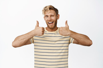 Smiling handsome man showing thumbs up and winking, say yes, praise and compliment, standing in t-shirt over white background