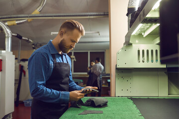 Shoe factory worker looking at pieces of new material. Side view of young man standing at cutting machine in workshop checking details cut out for future boots. Footwear manufacturing industry concept