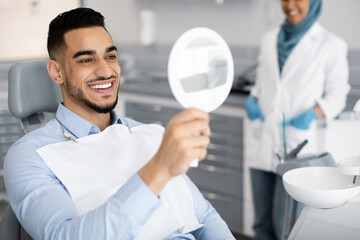 Happy Handsome Arab Man Looking To Mirror After Teeth Treatment In Clinic