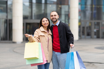 Happy international couple in casual wear posing with shopping bags, looking at camera and smiling near city mall