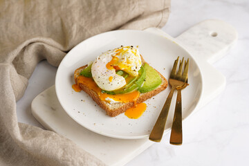 Savoury breakfast - A toast bread with cream cheese, avocado and poached egg on a white plate on marble tray