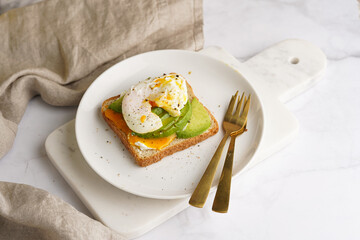 Savoury breakfast - A toast bread with cream cheese, avocado and poached egg on a white plate on marble tray