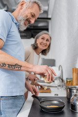 Smiling man holding spice while cooking pancake near blurred wife in kitchen.