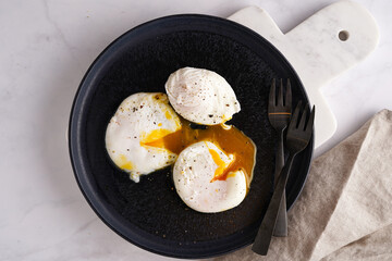 Three poached eggs with egg yolk on a black plate on a marble board and black forks